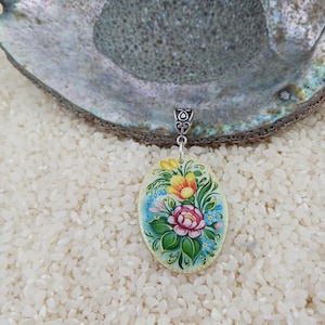 May include: Oval pendant with a hand-painted floral design. The pendant features a central pink rose surrounded by yellow, blue, and green flowers and leaves. The pendant is suspended from a silver-colored bail.