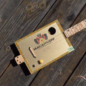 May include: A handmade cigar box guitar with a gold-colored box featuring the "MACANUDO" logo. The guitar has a wooden neck, strings, and a small yellow knob. The background is a weathered wooden surface.