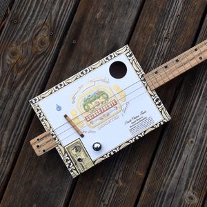 May include: A handmade cigar box guitar with a white and gold box featuring the "Arturo Fuente" logo. The guitar has a wooden neck, strings, and a small sound hole. The instrument rests on a weathered wooden surface.