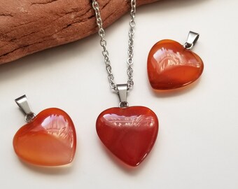 May include: Three heart-shaped pendants and necklaces. The pendants are a vibrant orange-red color, with a polished, glossy finish. The necklaces have a silver-colored chain and bail. The pendants are displayed on a white surface with a brown background.