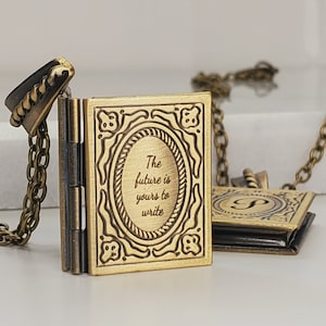 Peut inclure: Un collier pendentif en forme de livre de couleur bronze. Le pendentif en forme de livre ouvert présente un cadre ovale avec le texte "The future is yours to write". Un second pendentif avec la lettre "P" est visible en arrière-plan.