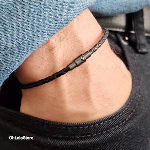 May include: Black braided leather bracelet with a gunmetal clasp. The bracelet is worn on a wrist, with a denim jacket and black trousers in the background. The OhLaLaStore logo is visible in the bottom left corner.