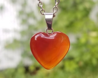 May include: A silver chain necklace with a heart-shaped carnelian pendant. The pendant is a deep orange color and has a smooth, polished finish.