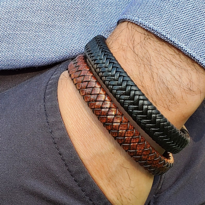 Pu&ograve; includere: Due braccialetti in pelle, uno nero e uno marrone, sono indossati al polso di una persona. I braccialetti sono intrecciati e hanno una trama tessuta.