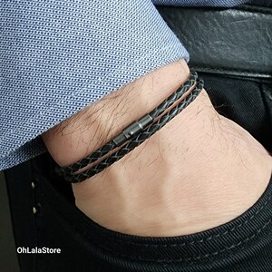 Peut inclure: Bracelet en cuir tressé noir avec fermoir magnétique. Le bracelet est enroulé autour du poignet. L'image montre le bracelet contre une chemise bleue à motifs et un pantalon noir. Le bracelet est un accessoire élégant.
