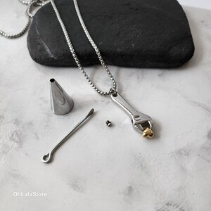 May include: A silver necklace with a wrench pendant and a gold heart charm. The necklace comes with a small funnel, a tool for inserting ashes, and a screw.