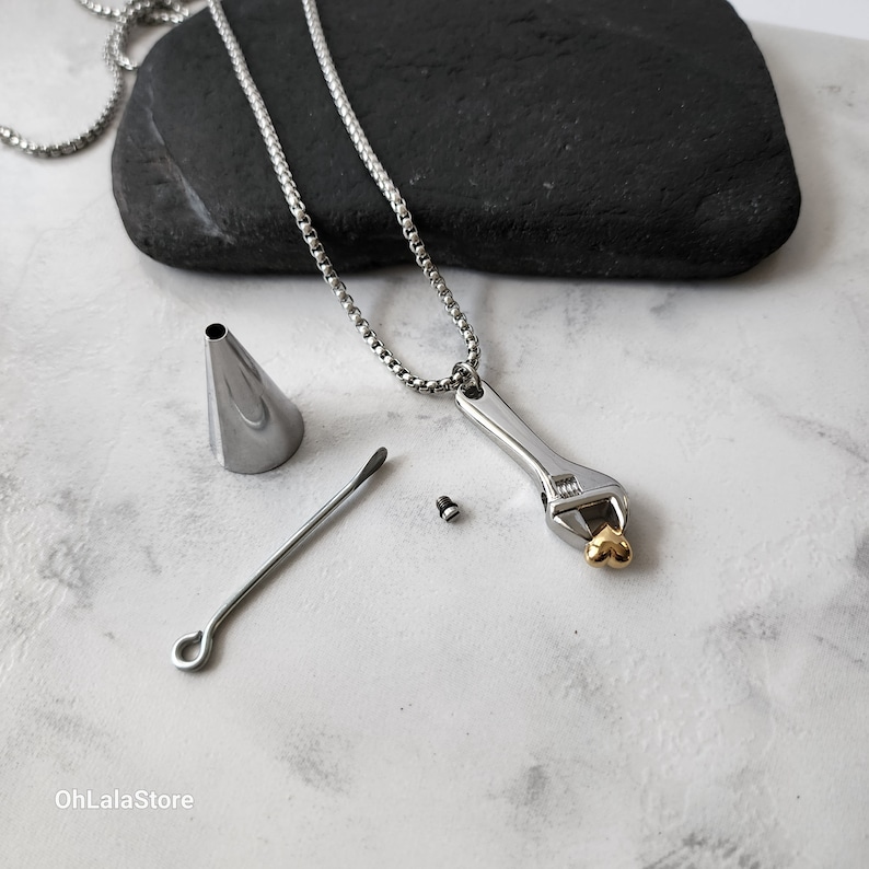 May include: A silver necklace with a wrench pendant and a gold heart charm. The necklace comes with a small funnel, a tool for inserting ashes, and a screw.