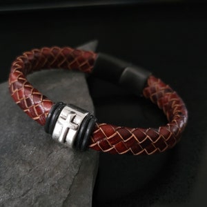 May include: A brown leather bracelet with a silver cross charm. The bracelet is braided and has a black rubber closure.