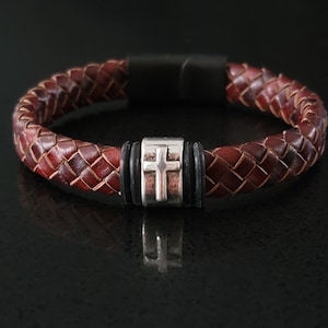 May include: Braided brown leather bracelet with a silver cross charm. The bracelet features a magnetic clasp and a woven design. The cross charm is centered on the bracelet.