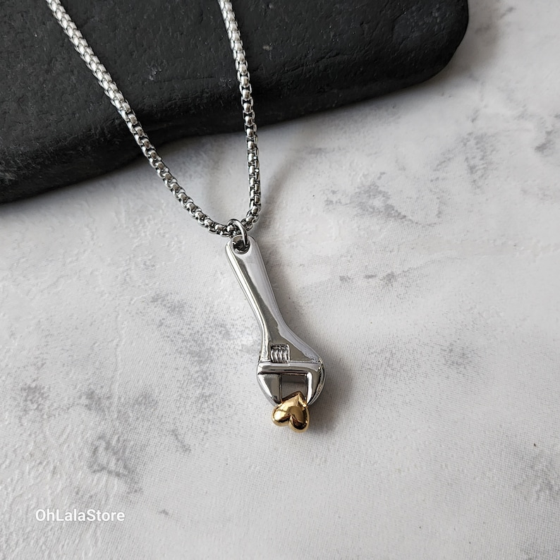 May include: A silver necklace with a silver wrench pendant holding a gold heart charm.