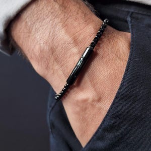 May include: A black bracelet featuring a cylindrical clasp and a beaded chain. The bracelet is crafted from a dark, possibly metallic material. The clasp is centrally positioned, suggesting a secure closure. The bracelet is worn on a wrist, with a dark blue shirt visible.