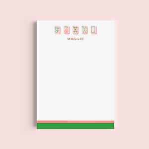 May include: A white notepad with a pink and green border. The notepad features a series of pink and green illustrated icons at the top, with the name "MAGGIE" printed in a light brown color. The notepad is rectangular and has a clean, minimalist design.