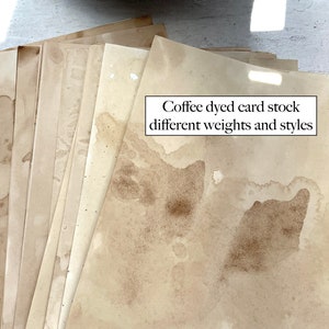 May include: A stack of coffee-dyed card stock in various shades of brown and beige. The paper has a mottled appearance with darker coffee stains and the text "Coffee dyed card stock different weights and styles".