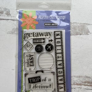 May include: Clear stamp set with travel-themed designs, including a bicycle, camera, airplane, and suitcase. Text includes "getaway," "take it easy," and "Trip of a Lifetime!" The packaging reads "Anyone Can Stamp!" and "HERO ARTS."