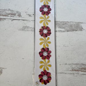 May include: A package of Sizzix decorative strip dies featuring a repeating pattern of yellow and burgundy flowers. The flowers have white centers. The package includes the text "Windmill Daisies" and "Sizzlits Decorative Strip".