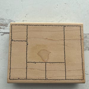 May include: A rectangular wooden stamp with a segmented design. The stamp features a series of interconnected rectangles and squares outlined in black ink. The wood has a natural, light brown color with a water stain.