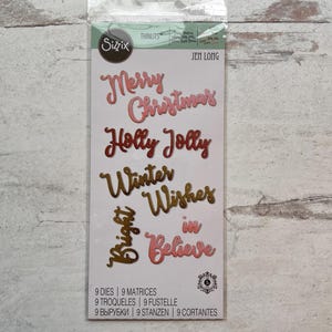 May include: A package of Sizzix Thinlits dies with Christmas-themed words in pink and gold. The words include "Merry Christmas," "Holly Jolly," "Winter Wishes," "Bright," and "Believe." The package also lists the number of dies and matrices.