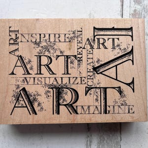 May include: Wooden art stamp with the word "ART" in large, bold letters, repeated multiple times with other words like "INSPIRE", "VISUALIZE", and "IMAGINE". The stamp has a light brown color and a rectangular shape.