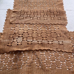 May include: A tan-colored, open-weave crochet textile with a floral pattern. The edges have a scalloped design. The textile appears to be handmade and is likely used as a decorative element, such as a doily or table runner.