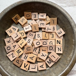May include: A collection of wooden Scrabble tiles with black letter printing, scattered inside a rustic, round metal bowl. The tiles display various letters and point values, perfect for word games or crafting projects.