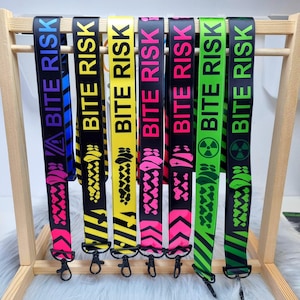 May include: A selection of lanyards, each printed with "BITE RISK" in different colours, such as blue, yellow, pink, and green. They have a black background and a metal clasp. The lanyards are displayed on a wooden rack.