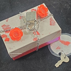 May include: A small silver padlock with a silver chain and two red rose charms on a white box with a red border. The box is sitting on a black surface.