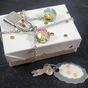 May include: A silver padlock with a chain attached to two iridescent fish charms. The charms are rainbow colored and have a 3D effect. The padlock and chain are on a white box with gold polka dots.
