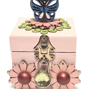 May include: A small, pink wooden box with a butterfly and flower design. The box has a metal clasp and a green gem lock. The butterfly is blue and the flowers are pink with maroon centers. The box is a decorative item.