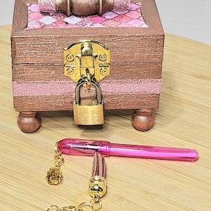 May include: A small wooden box with a pink and white patterned lid, a gold lock and key, and a pink pen with a gold charm attached. The box is sitting on a wooden surface.