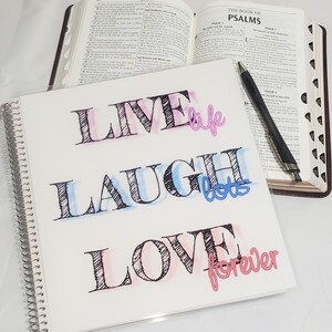 May include: A white journal with a clear plastic cover and a spiral binding. The journal has the words "Live life, Laugh lots, Love forever" written on it in black, pink, and blue ink.