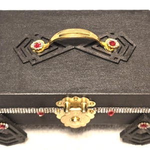 May include: A black decorative box with a gold handle and clasp. The box features red and clear crystal accents, with decorative corner pieces. The box is rectangular and has a textured surface.