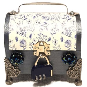 May include: A small, decorative chest with a floral pattern on the lid and a combination lock. The chest is primarily gray with silver accents and decorative corner pieces. The lid features a cream background with blue floral designs.