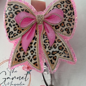 May include: A pink and leopard print bow-shaped badge reel with glitter accents. The bow features a pink center and leopard print wings. The badge reel has a metal clip and the text "The Garnet Magnolia" is visible.