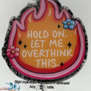 May include: A handmade badge reel with a flame design in orange, pink, and red. The text "HOLD ON. LET ME OVERTHINK THIS." is in the center, with flower and star accents. The edges are outlined in black glitter.