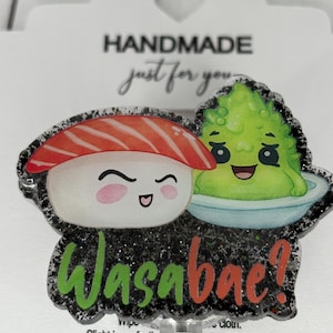 Puede incluir: Un amuleto de resina negra y plateada con purpurina, con un diseño de dibujos animados de sushi y wasabi. El sushi es rojo y blanco con una envoltura de alga negra. El wasabi es verde y tiene una cara linda. El amuleto dice "Wasabac!" en letras naranjas y verdes.