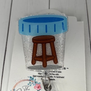 May include: A glittery badge reel featuring a brown stool inside a blue-lidded container. The badge reel has a clear plastic clip and a retractable cord. The Garnet Magnolia logo is visible at the bottom.