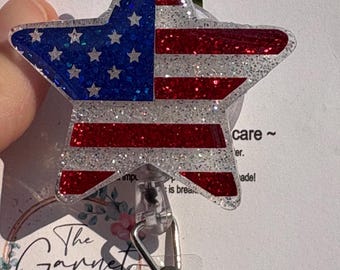 May include: A star-shaped badge reel featuring a glittery American flag design with a blue field of white stars on the left and red and white stripes on the right. The badge reel includes a clear plastic strap and a metal swivel clip attachment.