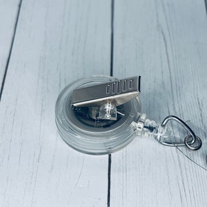 May include: A clear, round retractable badge reel with a silver metal clip and a silver metal pull. The reel has a clear plastic casing and a metal spring mechanism. The badge reel is designed to hold an ID badge or other small items.