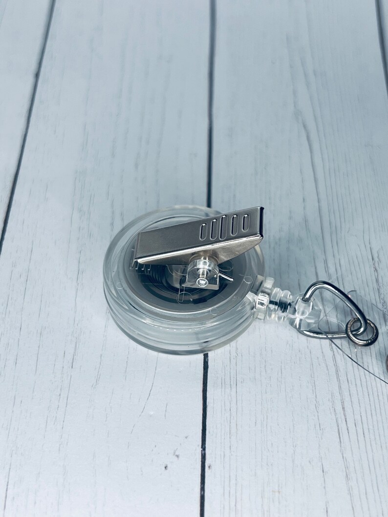 May include: A clear, round retractable badge reel with a silver metal clip and a silver metal pull. The reel has a clear plastic casing and a metal spring mechanism. The badge reel is designed to hold an ID badge or other small items.