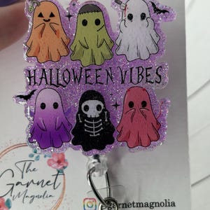 May include: A Halloween-themed badge reel featuring six cartoon ghosts in various colors, including orange, green, white, purple, black, and pink. The phrase "HALLOWEEN VIBES" is displayed across the center. The badge reel has a glittery purple border.