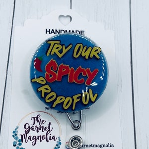 May include: A blue and yellow badge holder with the text "TRY OUR SPICY PROPOFOL" in red and yellow letters. A red chili pepper is on the left side of the badge holder. The badge holder is attached to a silver retractable badge reel.