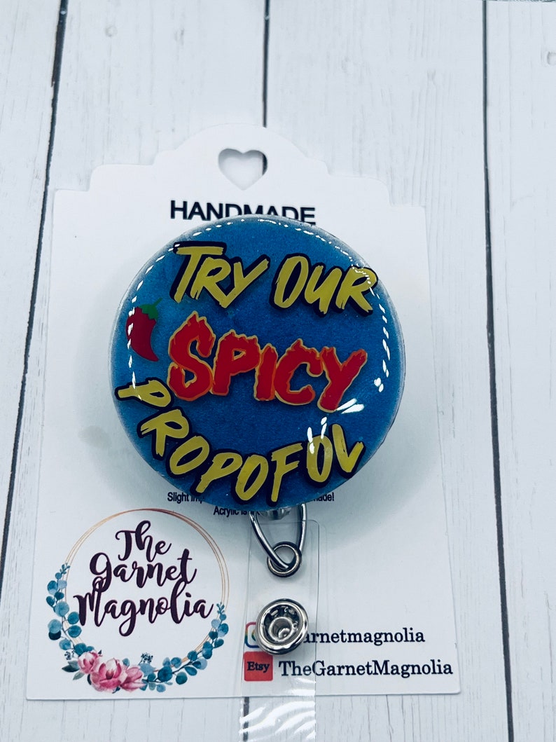 May include: A blue and yellow badge holder with the text "TRY OUR SPICY PROPOFOL" in red and yellow letters. A red chili pepper is on the left side of the badge holder. The badge holder is attached to a silver retractable badge reel.