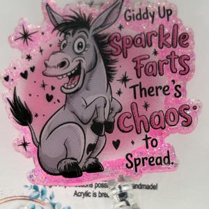 以下が含まれることがあります： ピンク色のキラキラ光るバッジ。漫画のロバのイラストが描かれています。バッジには「Giddy Up Sparkle Farts There's Chaos to Spread」という文字が書かれています。「HANDMADE」という文字が上部にあります。