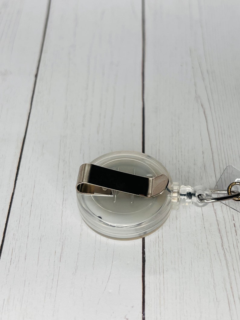 May include: A clear retractable badge holder with a silver clip. The holder is on a white wooden surface.