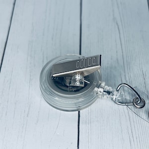 May include: A clear plastic retractable badge holder with a silver clip. The holder is on a white wooden surface.