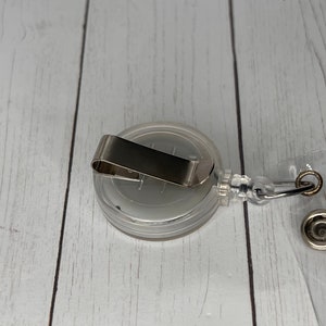 May include: A clear retractable badge holder with a silver clip and a clear plastic strap. The holder is designed to hold a badge or ID card.