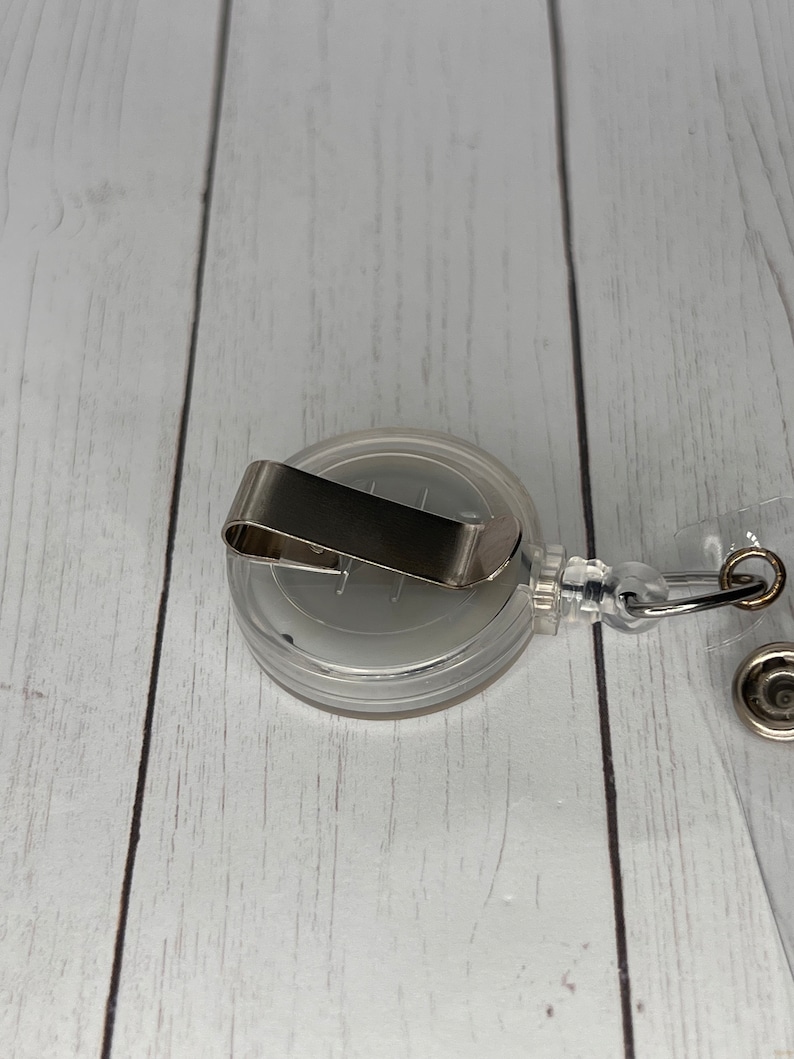 May include: A clear retractable badge holder with a silver clip and a clear plastic strap. The holder is designed to hold a badge or ID card.