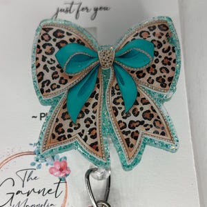 May include: A handmade badge reel with a bow design. The bow features a leopard print and teal accents, with glitter and a rhinestone centre. The badge reel has a retractable cord and a metal clip. The text "HANDMADE just for you" is visible.
