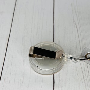 May include: A clear, round badge reel with a metal clip and a retractable cord. The reel has a silver-colored metal clip on the back and a clear plastic casing. The cord is attached to a clear plastic loop.