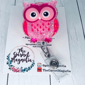 May include: Pink glitter owl charm with a clear plastic retractable badge holder. The charm is attached to the holder with a silver metal clip. The holder has a clear plastic strap and a silver metal button. The charm is attached to a white card with the text "The Garnet Magnolia" and social media handles.